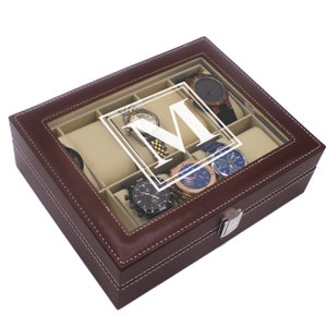 May include: A brown leather watch box with a clear glass top, featuring a white letter "M" and several watches. The box has a beige interior and white stitching around the edges. A silver clasp secures the box.