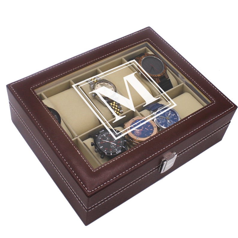 May include: A brown leather watch box with a clear glass top, featuring a white letter "M" and several watches. The box has a beige interior and white stitching around the edges. A silver clasp secures the box.