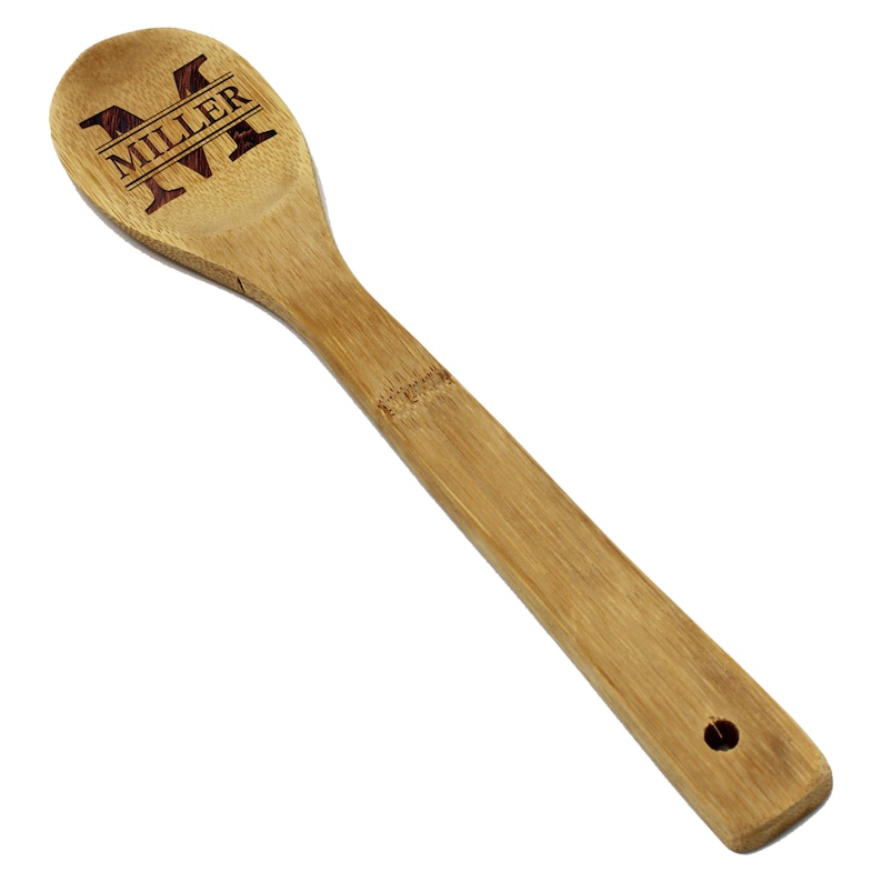May include: A bamboo wooden spoon with a long handle and a rounded head. The head of the spoon features the name "MILLER" and the letter "M" engraved in a dark brown color. The spoon has a hole at the end of the handle.