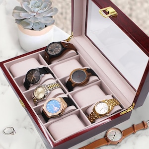 May include: A dark cherry wood watch display case with a glass lid and gold-tone metal hinges. The interior features six beige velvet-lined compartments, each containing a watch with various band styles, including leather, metal, and wood links.