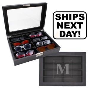 May include: A black rectangular eyewear storage case with a clear glass lid and eight interior compartments. The case features a textured exterior finish and a silver-toned metal latch. One view shows the case open with various sunglasses inside.