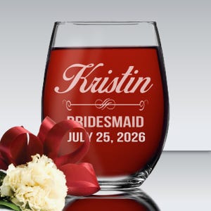 May include: A clear stemless wine glass filled with red liquid, featuring white etched text that reads Kristin, BRIDESMAID, and JULY 25, 2026, with a decorative flourish line. A white carnation with a dark red ribbon is positioned in the foreground.