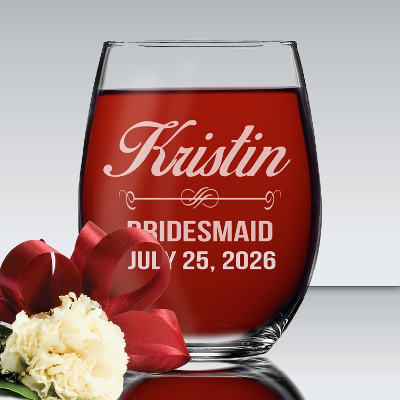 May include: A clear stemless wine glass filled with red liquid, featuring white etched text that reads Kristin, BRIDESMAID, and JULY 25, 2026, with a decorative flourish line. A white carnation with a dark red ribbon is positioned in the foreground.