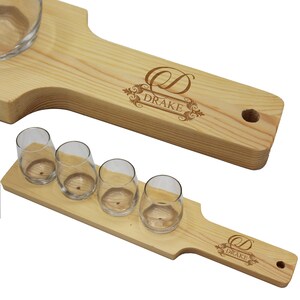 K&ouml;nnte beinhalten: Zwei Holzservierbretter mit Schnapsgl&auml;sern aus klarem Glas. Die Bretter sind aus hellem Holz gefertigt und tragen das eingravierte Wort "DRAKE" mit einem dekorativen Design. Ein Brett hat eine runde Sch&uuml;ssel zum Dippen.