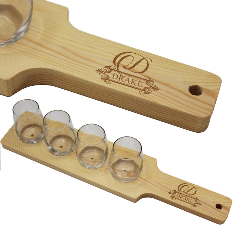 K&ouml;nnte beinhalten: Zwei Holzservierbretter mit Schnapsgl&auml;sern aus klarem Glas. Die Bretter sind aus hellem Holz gefertigt und tragen das eingravierte Wort "DRAKE" mit einem dekorativen Design. Ein Brett hat eine runde Sch&uuml;ssel zum Dippen.