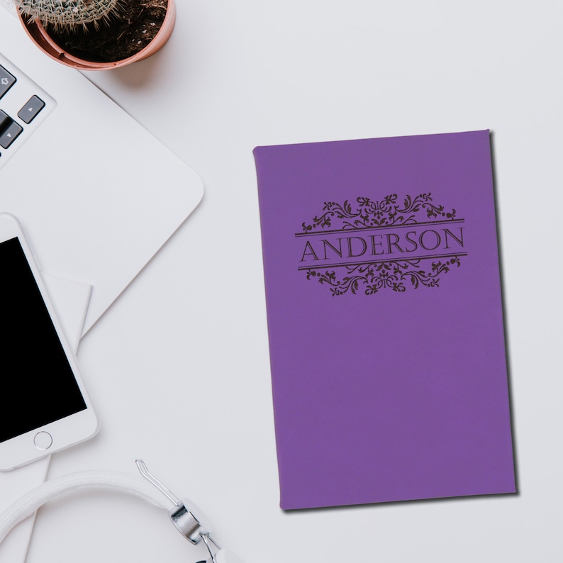 May include: A purple journal with the name "ANDERSON" in a decorative font. The journal has an ornate border design. The journal is on a white surface, next to a laptop, a smartphone, and headphones.