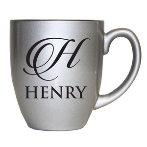 Pu&ograve; includere: Una tazza in ceramica color argento metallizzato con una grande lettera H calligrafica nera nella parte superiore e il nome HENRY stampato in un carattere nero in grassetto con grazie direttamente sotto la lettera.