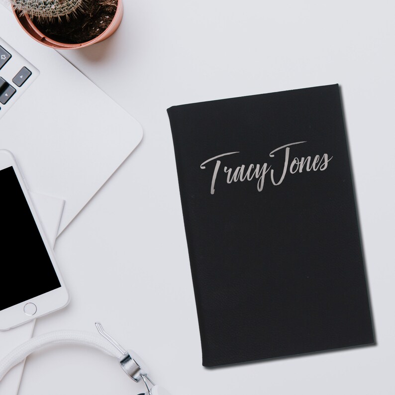May include: Black notebook with the name "Tracy Jones" in silver script. The notebook is on a white surface, next to a laptop, a smartphone, and headphones. A small potted cactus is also visible.