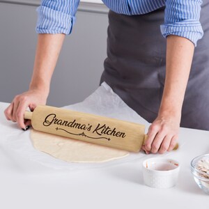 Puede incluir: Un rodillo de madera de color claro con las palabras "Grandma's Kitchen" en una fuente decorativa. El rodillo se utiliza para aplanar la masa sobre una superficie blanca. Una persona est&aacute; usando el rodillo.