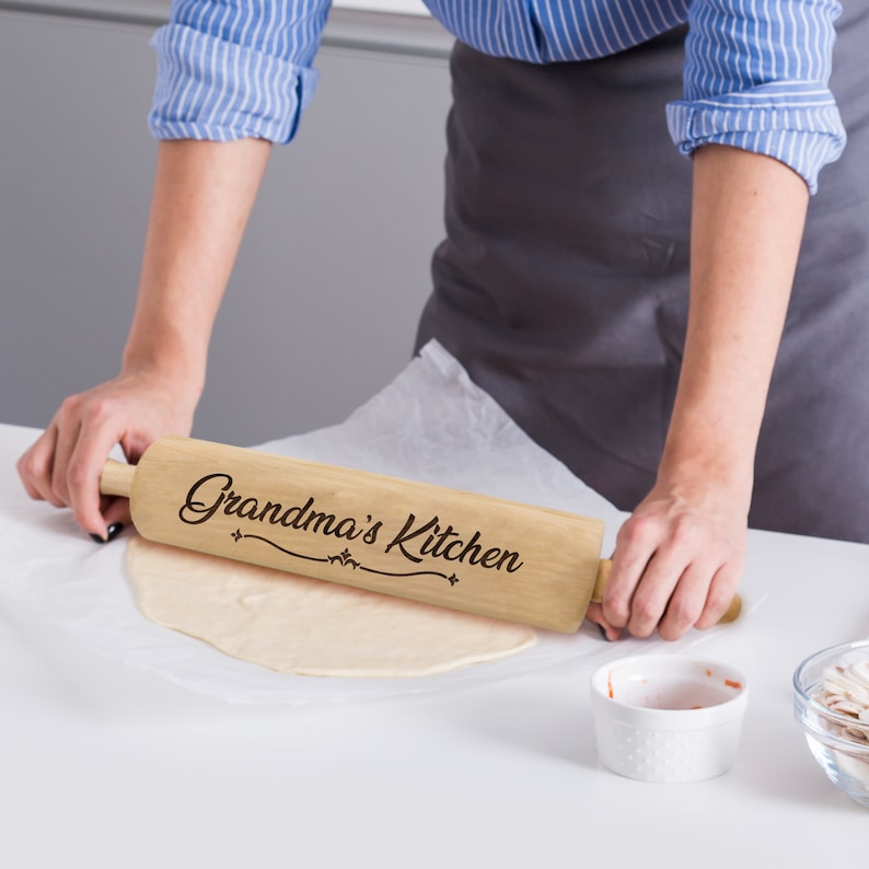 Puede incluir: Un rodillo de madera de color claro con las palabras "Grandma's Kitchen" en una fuente decorativa. El rodillo se utiliza para aplanar la masa sobre una superficie blanca. Una persona est&aacute; usando el rodillo.