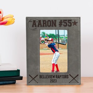 May include: Gray leatherette photo frame with a baseball graphic and the text "AARON #55*" at the top. The frame has a photo of a young boy in a baseball uniform holding a bat. The bottom of the frame has the text "BELLEVUE RAPTOR 2023" and two crossed baseball bats.