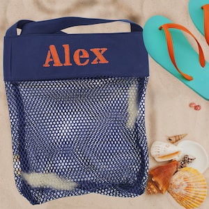 May include: A blue mesh beach bag with a navy blue top flap and the name "Alex" embroidered in orange. The bag is sitting on a sandy surface with seashells.