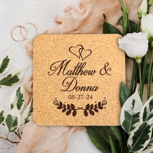 May include: Cork coaster with the text "Matthew & Donna 08-27-24" and two intertwined hearts above the names. The text is surrounded by a decorative leafy border.