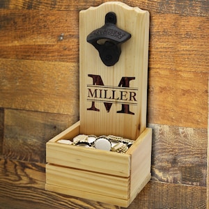 May include: Wooden wall-mounted bottle opener with a built-in cap catcher. The opener is black and mounted on a light-colored wooden board. The board has the name "MILLER" engraved in a dark brown color. The cap catcher is a small wooden box filled with bottle caps.