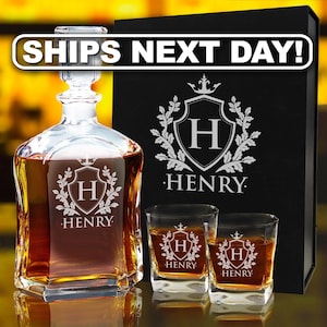 Puede incluir: Un decantador de cristal transparente y dos vasos cuadrados a juego llenos de líquido ámbar. Cada pieza presenta un escudo grabado con la letra H y el nombre HENRY. Una caja de presentación negra con el mismo escudo y nombre se sitúa detrás.