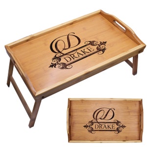 May include: A rectangular bamboo breakfast tray with foldable legs and integrated side handles. The surface features a dark laser-engraved design with a large letter D and the name DRAKE centered within an ornate decorative frame.