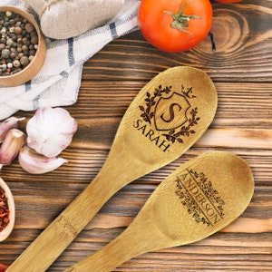 May include: Two wooden spoons with engraved designs. One spoon features the name "SARAH" with a shield and initial "S". The other spoon has the name "ANDERSON" with floral accents. The spoons are on a wooden surface.