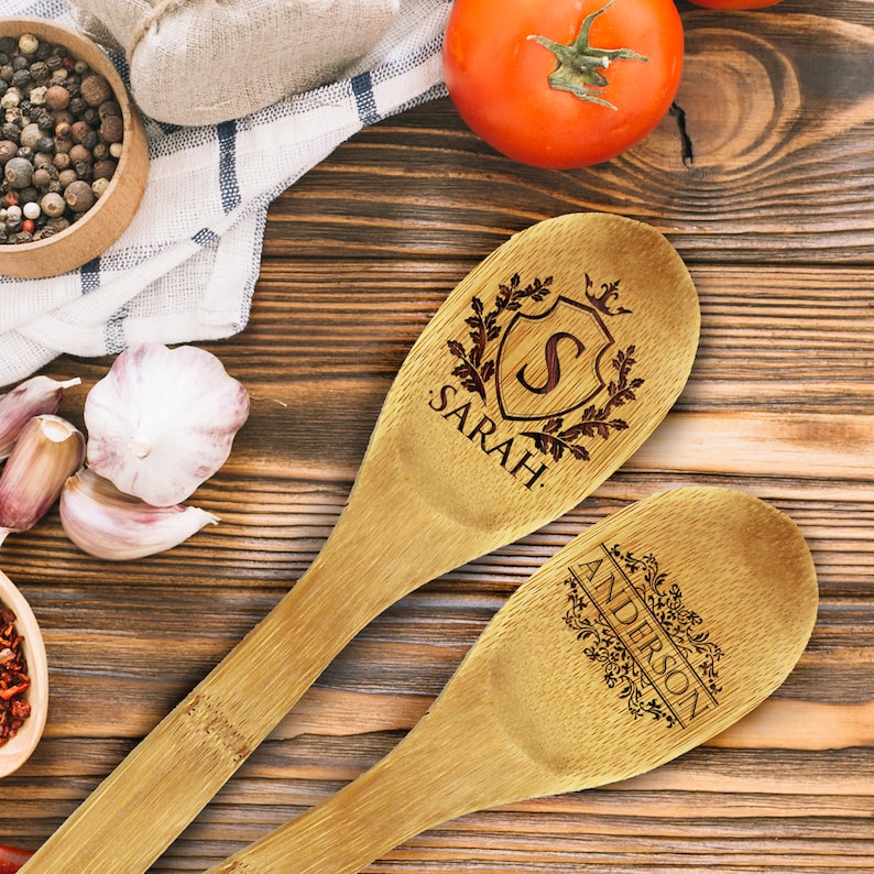 May include: Two wooden spoons with engraved designs. One spoon features the name "SARAH" with a shield and initial "S". The other spoon has the name "ANDERSON" with floral accents. The spoons are on a wooden surface.