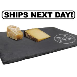 May include: A rectangular black slate serving board featuring a circular engraved monogram with the letter M and the name MILLER. Two pieces of cheese are placed on the surface. The text SHIPS NEXT DAY! is displayed in a white box above the board.