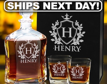 Pode incluir: Um decanter de vidro transparente e dois copos quadrados a condizer, cheios de líquido âmbar. Cada peça apresenta um brasão gravado com a letra H e o nome HENRY. Uma caixa de apresentação preta com o mesmo brasão e nome está atrás.