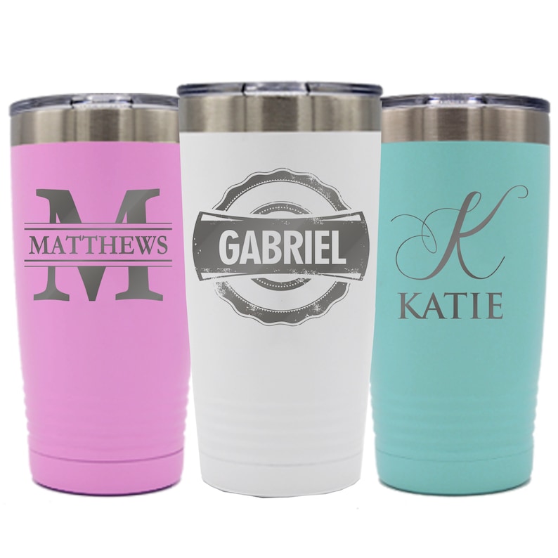 May include: Three stainless steel tumblers with personalized names. The tumbler on the left is pink with the name "MATTHEWS" and the initial "M". The middle tumbler is white with the name "GABRIEL" in a vintage style logo. The tumbler on the right is turquoise with the name "KATIE" and the initial "K".