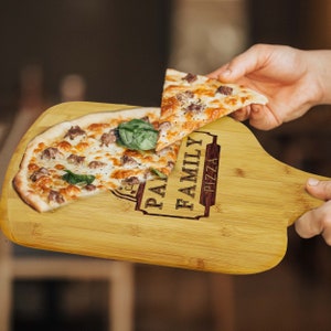 May include: A bamboo pizza peel with a whole pizza and a slice being held. The pizza has sausage, cheese, and basil. The peel has the words "FAMILY PIZZA" engraved on it.