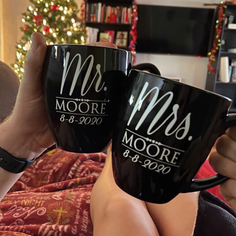 May include: Two black ceramic mugs, one inscribed "Mr. MOORE 8-8-2020" and the other "Mrs. MOORE 8-8-2020". The mugs are held up, with a Christmas tree and a television in the background. The mugs are a wedding or anniversary gift.