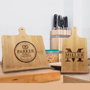 May include: Two bamboo recipe holders with engraved text. One reads "Parker Family Recipes Fresh & Delicious" in a circular design. The other displays the name "Miller" with a large initial "M". Knives are in a wooden block.