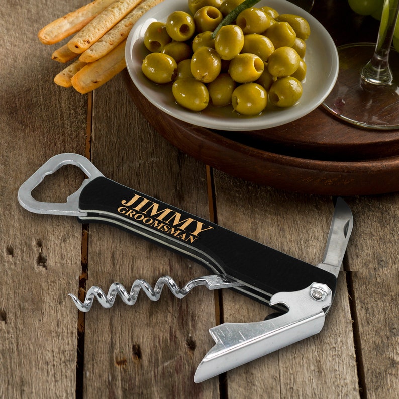 Op de afbeelding: Een zwarte multitool met een kurkentrekker, een flesopener en een klein mes. De tool heeft een zwarte handgreep met de tekst "JIMMY GROOMSMAN" in gouden letters.
