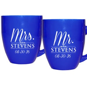 May include: Two cobalt blue mugs with white text. One mug reads "Mrs. STEVENS 08-30-26" and the other reads "Mr. STEVENS 08-30-26". The mugs have a curved shape and a handle.
