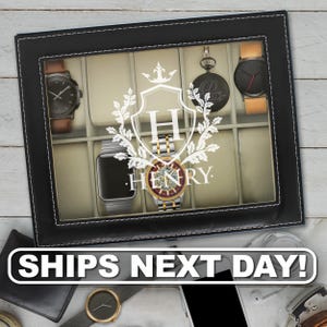 May include: A black rectangular watch display case with a glass lid featuring a white printed crest with the letter H and the name HENRY. The interior contains beige velvet cushions holding various wristwatches and a pocket watch.
