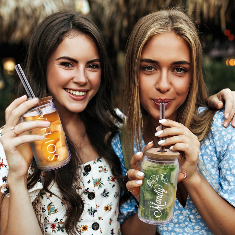 Puede incluir: Dos amigas disfrutando de bebidas en vasos de pl&aacute;stico transparente personalizados con pajitas. Los vasos tienen los nombres "Sophia" y "Mandy" escritos en ellos. Los vasos est&aacute;n decorados con un dise&ntilde;o de hojas verdes.