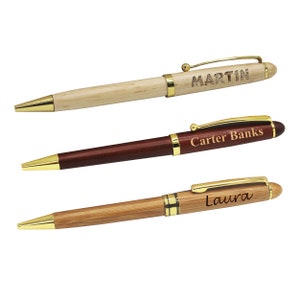 May include: Three personalized pens with gold accents. One is light wood with "MARTIN" engraved, another is dark brown with "Carter Banks", and the third is bamboo with "Laura".
