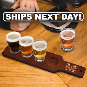 May include: A dark brown wooden beer flight paddle with four circular cutouts for glasses. Three glasses filled with different shades of beer sit in the paddle, while one glass sits separately. The paddle features the engraved letter M and MILLER.