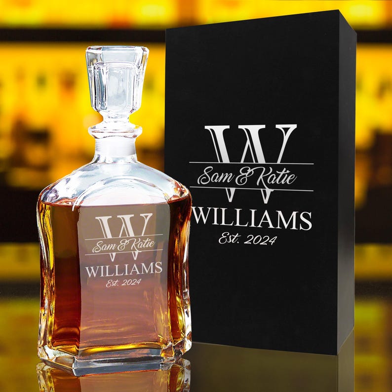 May include: A clear glass decanter filled with amber liquid, featuring a square stopper and an etched design with a large letter W, the names Sam & Katie, the surname WILLIAMS, and Est. 2024. Beside it stands a black rectangular gift box.