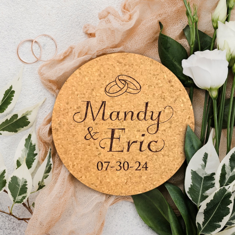 May include: A round cork coaster with the names Mandy & Eric and the date 07-30-24 engraved in brown lettering. Two gold rings are depicted above the names.