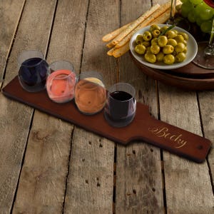 May include: A wooden wine flight paddle with four glasses filled with different colored wines. The paddle has a handle and the name "Becky" engraved on it. Olives, breadsticks, and grapes are in the background.