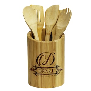 May include: A bamboo utensil holder with a personalized engraved design. The holder features a monogrammed initial "D" with the name "Drake" and the date "Est. 07.29.17". The holder is filled with four wooden utensils.