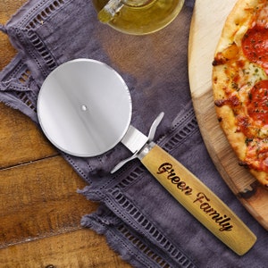 May include: A stainless steel pizza cutter with a wooden handle engraved with "Green Family" rests on a purple cloth. A partially visible pizza with pepperoni sits on a wooden cutting board, with a bottle of oil in the background.