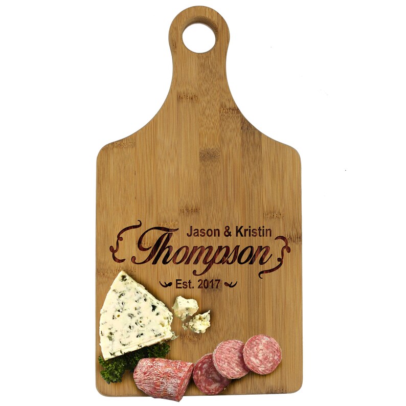 May include: A personalized bamboo cutting board with a handle. The board is engraved with the text "Jason & Kristin Thompson's Est. 2017". The board has a piece of blue cheese and slices of salami on it.