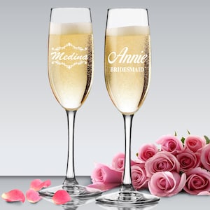 May include: Two champagne flutes with personalized engravings. The flute on the left says "Medina" and the flute on the right says "Annie Bridesmaid". The flutes are filled with champagne and are surrounded by pink roses.
