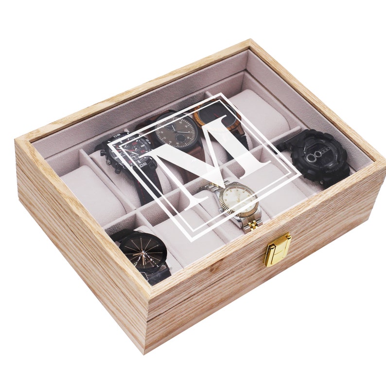 May include: A rectangular light-colored wooden watch box with a glass lid featuring a white letter M inside a square frame. The interior is lined with light gray fabric and contains multiple compartments, some holding various wristwatches.