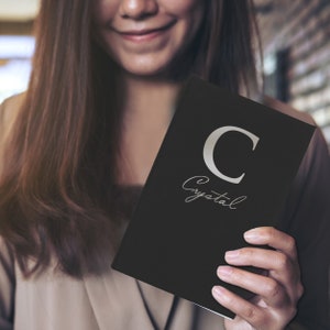Puede incluir: Un cuaderno negro con la letra "C" plateada y el nombre "Crystal" en escritura cursiva. El cuaderno lo sostiene una persona con cabello largo casta&ntilde;o.