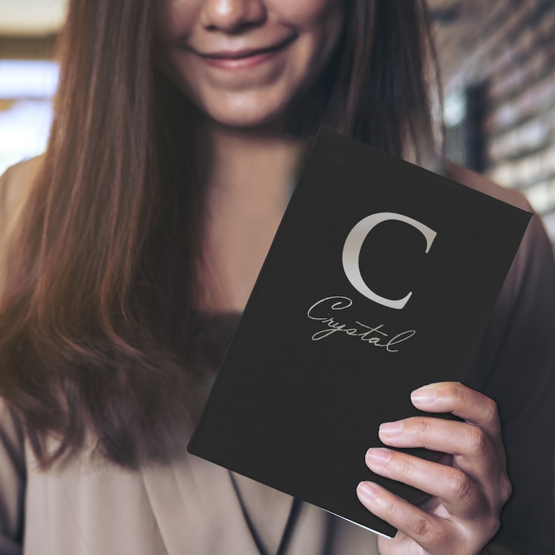 Puede incluir: Un cuaderno negro con la letra "C" plateada y el nombre "Crystal" en escritura cursiva. El cuaderno lo sostiene una persona con cabello largo casta&ntilde;o.