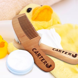 May include: Two wooden baby combs with the name "Carter's" engraved on them. The combs are on a yellow towel with a stuffed duck toy.