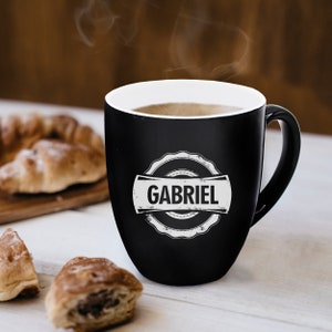 Pu&ograve; includere: Una tazza in ceramica nera opaca con interno bianco e manico. La parte anteriore presenta un emblema circolare bianco con bordo smerlato contenente il nome GABRIEL in grassetto maiuscolo. Il vapore sale dal liquido caldo nella tazza.