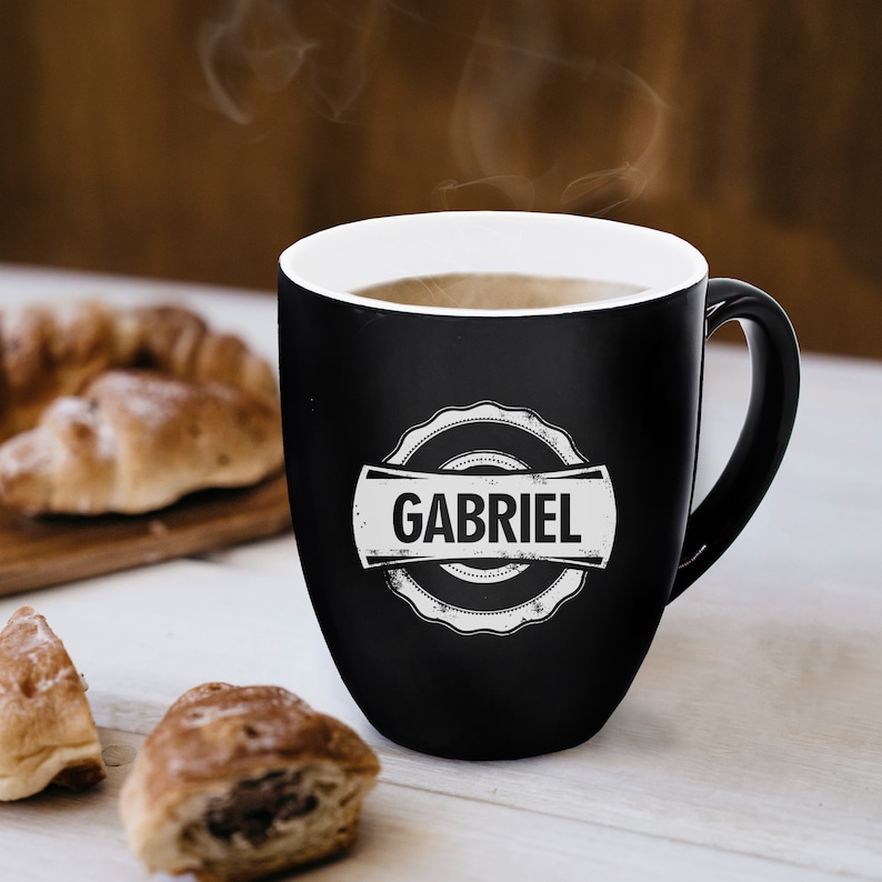 Pu&ograve; includere: Una tazza in ceramica nera opaca con interno bianco e manico. La parte anteriore presenta un emblema circolare bianco con bordo smerlato contenente il nome GABRIEL in grassetto maiuscolo. Il vapore sale dal liquido caldo nella tazza.
