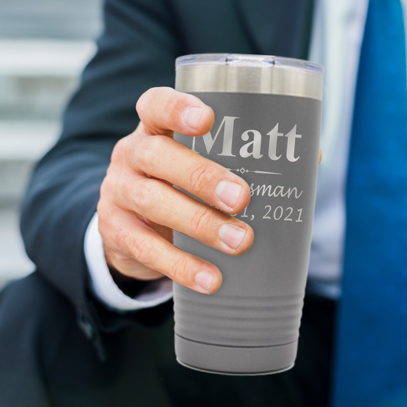 May include: Gray stainless steel tumbler with a silver lid. The tumbler has the name "Matt" engraved on it, along with the words "Best Man" and the date "1, 2021".