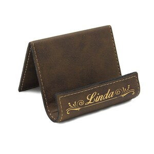 May include: A brown faux leather phone stand with a gold inscription that reads "Linda" with a decorative flourish.
