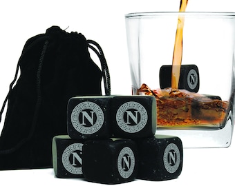 May include: Six black square whiskey stones with a silver letter "N" in a circle design. The stones are in a black velvet bag and are being used to chill a glass of whiskey.